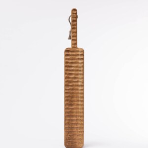 May include: A long, wooden serving board with a textured surface and a rope handle. The board is light brown in color.