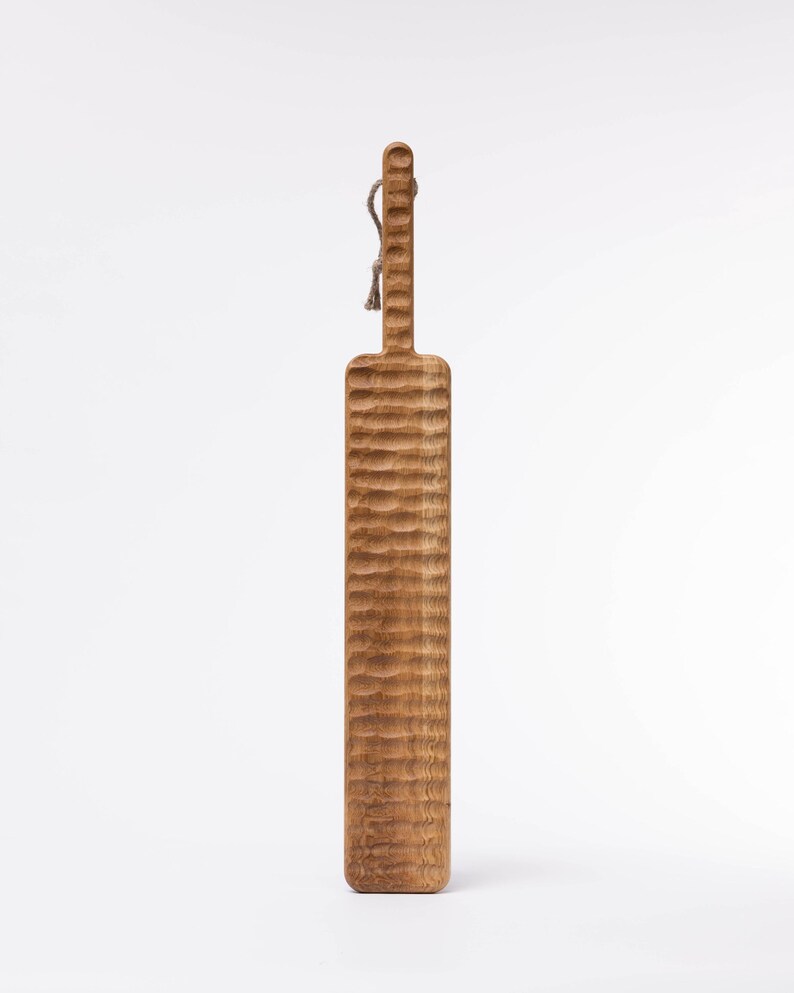 May include: A long, wooden serving board with a textured surface and a rope handle. The board is light brown in color.