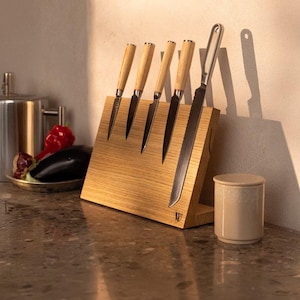 May include: A wooden magnetic knife block with five knives, including a bread knife, attached to it. The block is light brown and has a W2 logo on the bottom. The knives have wooden handles and are arranged in a row.