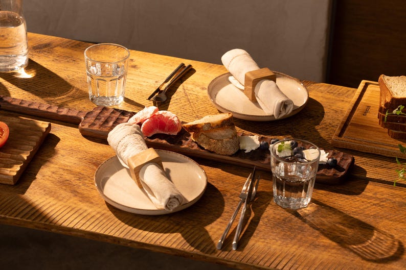 May include: A wooden serving tray with a light brown finish, featuring a napkin ring, a slice of grapefruit, a piece of bread, and a small bowl of blueberries. The tray is set on a wooden table with two glasses of water and a set of silverware.
