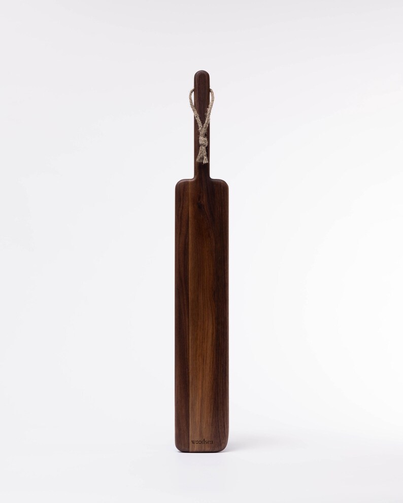 May include: A long, rectangular wooden cutting board with a dark brown finish. It has a rope handle and the word "woodlot" is engraved on the bottom.
