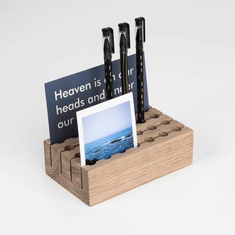 May include: A wooden desk organiser with a light brown finish. The organiser has multiple slots for pens, pencils, and other office supplies. There are three black pens with gold accents in the organiser. A blue card with white text that reads "Heaven is in our heads and not our" is in the organiser. A photo of a body of water with a rocky shore is in the organiser.