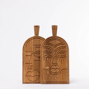 May include: Two wooden cutting boards with carved faces. The boards are made of light brown wood and feature stylized faces with geometric designs. Each board has a handle at the top. The faces have detailed eyes, noses, and mouths.