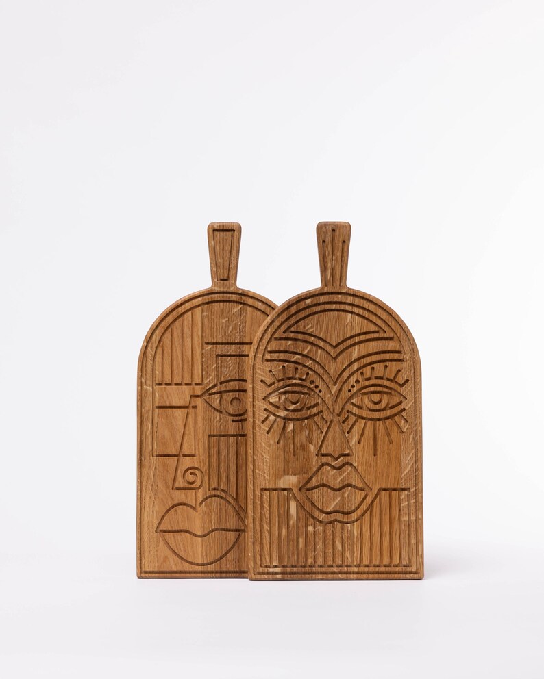 May include: Two wooden cutting boards with carved faces. The boards are made of light brown wood and feature stylized faces with geometric designs. Each board has a handle at the top. The faces have detailed eyes, noses, and mouths.