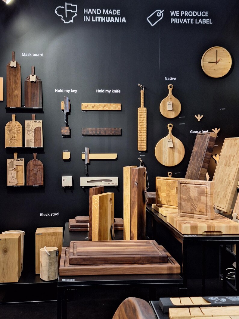 May include: A display of wooden cutting boards and serving trays, with various sizes and shapes. The boards are made in Lithuania and are labeled with names like "Native", "Hold my knife", and "Goose feet". The sign above reads "Hand Made in Lithuania" and "We Produce Private Label".