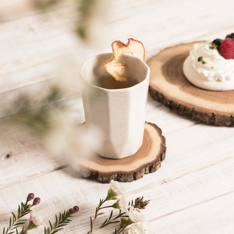 Puede incluir: Una taza de cer&aacute;mica blanca con forma hexagonal se encuentra sobre un posavasos de madera. La taza est&aacute; llena de una bebida de color claro y una rodaja de manzana seca flota en la parte superior. La taza est&aacute; sobre una superficie de madera blanca.
