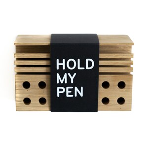 May include: A wooden pen holder with a black label that says "HOLD MY PEN". The holder has a series of holes for storing pens.