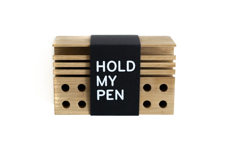 May include: A wooden pen holder with a black label that says "HOLD MY PEN". The holder has a series of holes for storing pens.