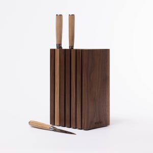 May include: A wooden knife block with a walnut finish and a striped design. The block has two knives with wooden handles inserted into it. A third knife with a wooden handle is lying on a white surface in front of the block.