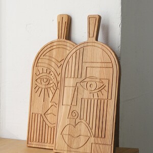 May include: Two wooden cutting boards with carved geometric designs resembling faces. Each board has a unique design with an eye, nose, and mouth.
