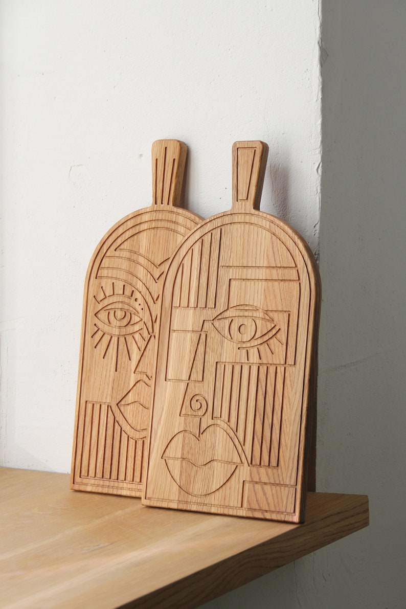 May include: Two wooden cutting boards with carved geometric designs resembling faces. Each board has a unique design with an eye, nose, and mouth.