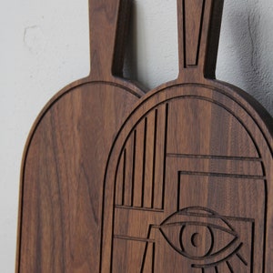 May include: Two wooden cutting boards, one with a geometric design and the other plain. The geometric design features a stylized eye and abstract shapes.