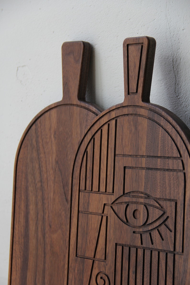 May include: Two wooden cutting boards, one with a geometric design and the other plain. The geometric design features a stylized eye and abstract shapes.