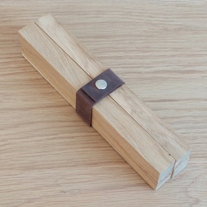 Può includere: Un supporto pieghevole in legno con un cinturino in pelle marrone e un bottone in metallo. Il supporto è in legno chiaro e ha una finitura naturale.
