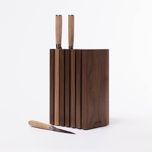 May include: A rectangular dark brown wooden knife block with vertical slats and two knives inserted into the top. A third knife with a light brown wooden handle and a silver blade lies horizontally in front of the block. The brand name is visible.