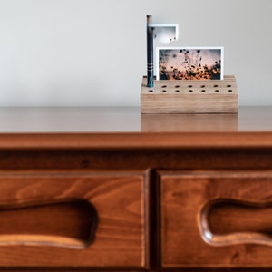 May include: A wooden photo holder with a natural finish and a grid of holes to hold photos. A photo of a field of flowers at sunset is displayed in the holder.