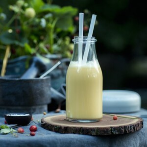 Puede incluir: Una botella de vidrio con un batido amarillo en su interior, dos pajitas blancas que sobresalen de la parte superior y un posavasos de madera debajo. La botella est&aacute; sobre un mantel azul con hojas verdes y bayas rojas esparcidas a su alrededor.