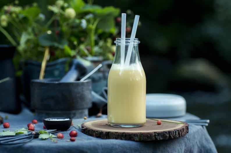 Puede incluir: Una botella de vidrio con un batido amarillo en su interior, dos pajitas blancas que sobresalen de la parte superior y un posavasos de madera debajo. La botella est&aacute; sobre un mantel azul con hojas verdes y bayas rojas esparcidas a su alrededor.
