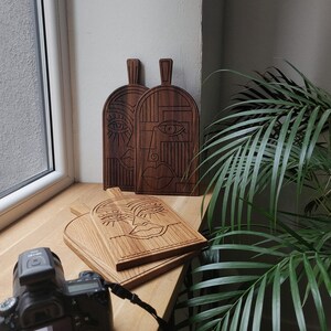 May include: Three wooden cutting boards with abstract face designs carved into them. The boards are brown and have a natural wood finish. The faces are stylized and have large eyes and lips.