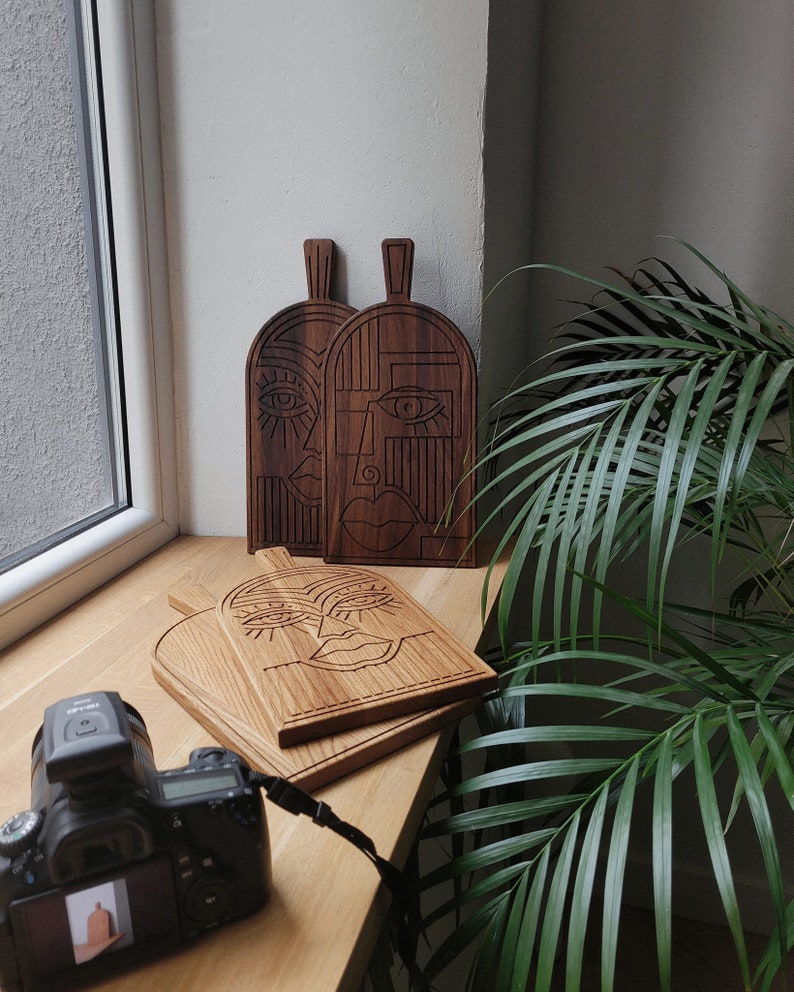 May include: Three wooden cutting boards with abstract face designs carved into them. The boards are brown and have a natural wood finish. The faces are stylized and have large eyes and lips.