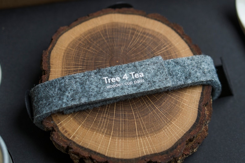 Puede incluir: Un juego de dos posavasos redondos de madera con una banda de fieltro gris. La banda tiene el texto "Tree 4 Tea wooden cup pads" impreso en ella.