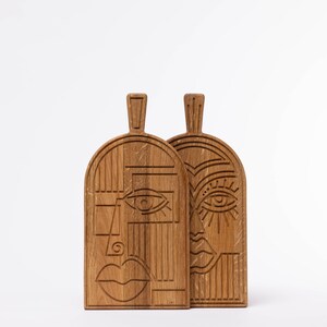 May include: Two wooden cutting boards with carved abstract face designs. The boards are made of light brown wood and feature a handle at the top. One face has a simple eye and mouth, while the other has more detailed features.