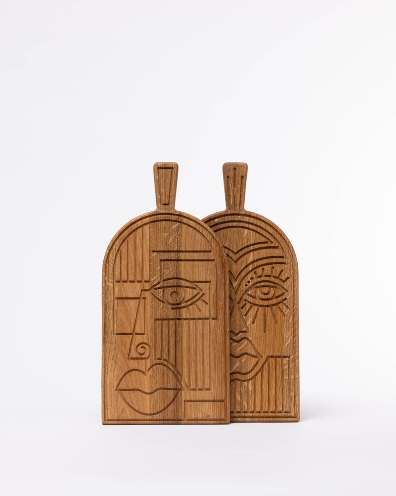 May include: Two wooden cutting boards with carved abstract face designs. The boards are made of light brown wood and feature a handle at the top. One face has a simple eye and mouth, while the other has more detailed features.