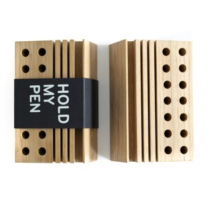 May include: Two wooden pen holders with a black label that reads "HOLD MY PEN". Each holder has multiple rows of holes for pens.