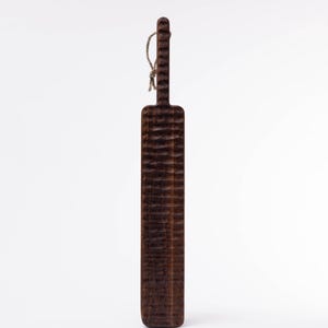 May include: A long, dark brown wooden cutting board with a textured surface and a rope handle.