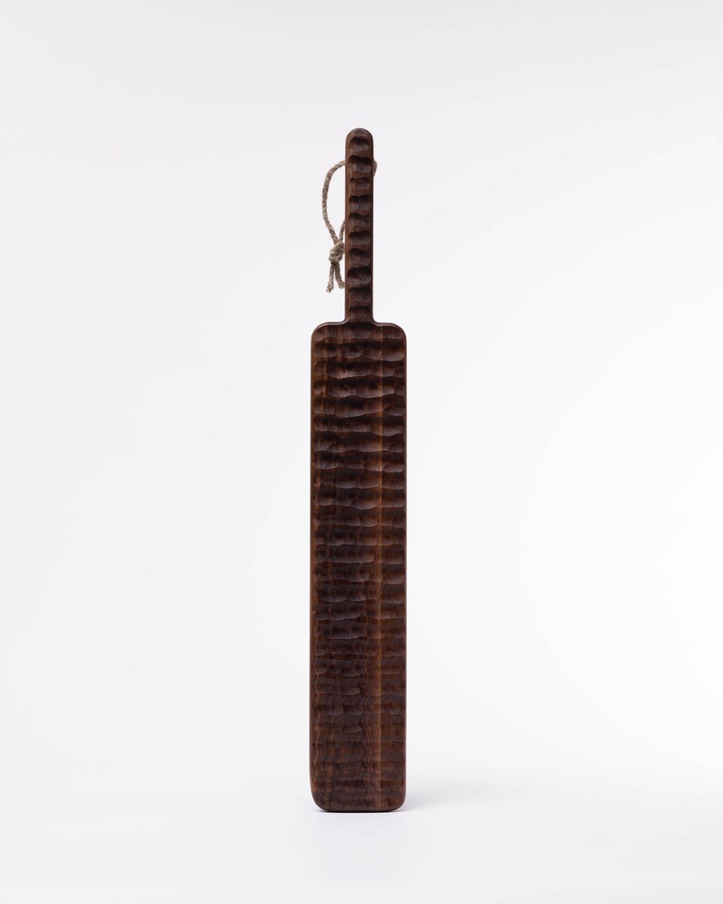 May include: A long, dark brown wooden cutting board with a textured surface and a rope handle.