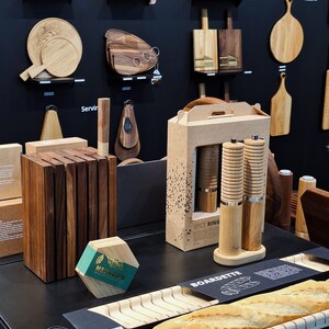 May include: A display of wooden kitchenware including a dark wood knife block, a hexagonal box labeled HEXAGON, a cardboard box labeled SPICE RING containing two wooden pepper mills, and a light wood baguette board labeled BOARDETTE.