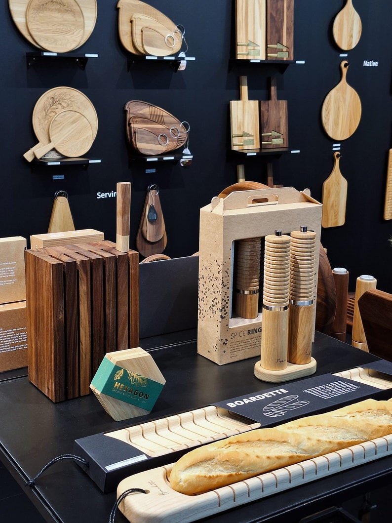 May include: A display of wooden kitchenware including a dark wood knife block, a hexagonal box labeled HEXAGON, a cardboard box labeled SPICE RING containing two wooden pepper mills, and a light wood baguette board labeled BOARDETTE.