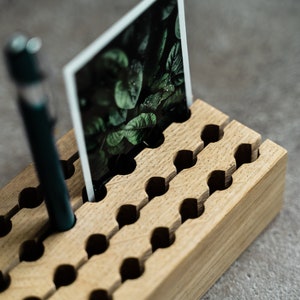 May include: A wooden organiser with slots for holding pens and photos. The organiser is made of light brown wood and has a natural finish. A green photo and a blue pen are in the organiser.