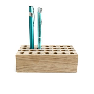 May include: A wooden pen holder with 36 holes, featuring two teal pens inserted. The holder is made of light brown wood with a natural grain.