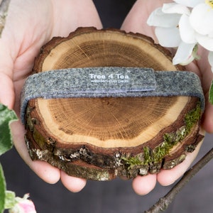 Puede incluir: Dos posavasos de madera hechos de corteza de &aacute;rbol con una banda de fieltro gris alrededor de la parte superior. La banda tiene el texto "Tree 4 Tea wooden cup pads" impreso en ella.