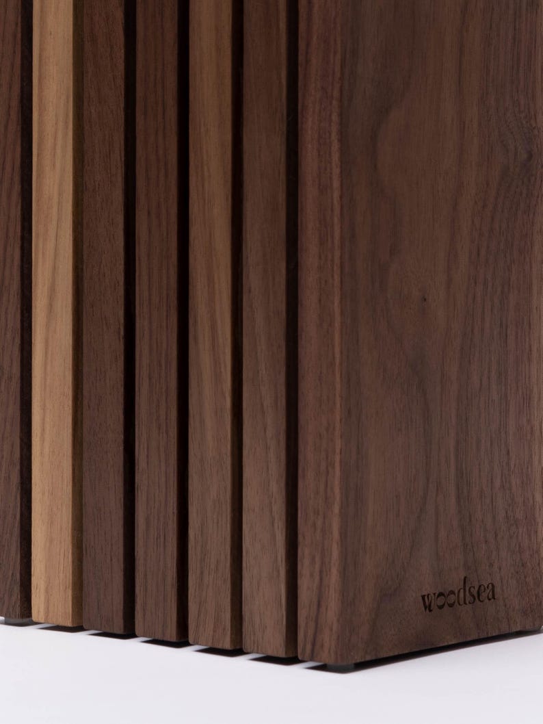 May include: A vertical wooden knife block constructed from multiple dark brown walnut slats with one lighter tan slat integrated into the design. The block features a smooth finish and the word woodsea is embossed on the lower right side.