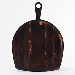 May include: Dark brown wooden cutting board with a handle and a circular hole. The board has a rounded shape and a natural wood grain pattern. The handle is attached to the top of the board.