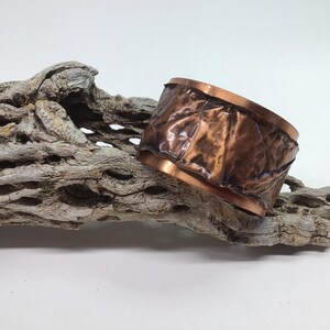May include: A handcrafted copper cuff bracelet with a textured, hammered design. The bracelet has a rich, reddish-brown color and is approximately 2 inches wide. It rests on a piece of weathered, light-colored wood, creating a rustic aesthetic.