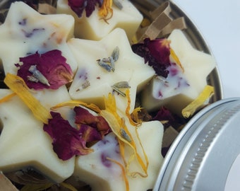 May include: A tin container filled with star-shaped wax melts. The melts are white and have dried flower petals and herbs embedded in them. The tin has a silver lid.