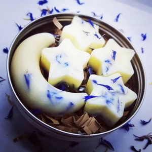May include: A set of five white wax melts in the shape of a crescent moon and stars. The wax melts are decorated with blue flower petals and are packaged in a silver tin.