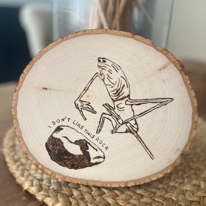 Puede incluir: Una rodaja de madera redonda con un diseño pirograbado. El diseño presenta una figura similar a una criatura y una roca, con el texto "I DON'T LIKE THIS ROCK". La madera tiene una veta natural y una estética rústica.