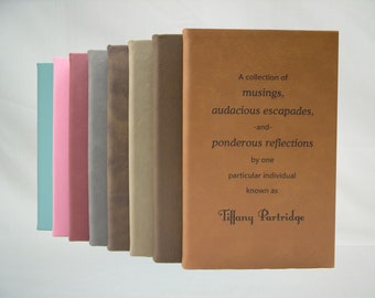 May include: A collection of ten leather-bound journals in various colors, including teal, pink, red, gray, brown, and beige. The front cover of the brown journal has the text "A collection of musings, audacious escapades, -and- ponderous reflections by one particular individual known as Tiffany Partridge".