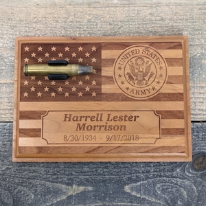 May include: A wooden plaque with an American flag design, a bullet casing holder, and a personalized inscription that reads "Harrell Lester Morrison 8/30/1934 - 9/17/2018". The plaque also features a United States Army seal.