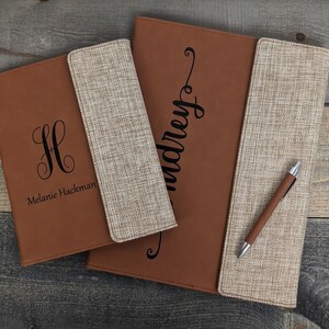 May include: Two brown leather and beige fabric personalized folders with a pen. The smaller folder has the monogram "H" and the name "Melanie Hackman". The larger folder has the name "Audrey" in cursive.