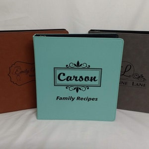 May include: Three personalized binders in brown, teal, and gray. The teal binder has the name "Carson" and the text "Family Recipes". The brown binder has the name "Emily C" and the gray binder has the name "Line Lane".