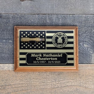 May include: A wooden plaque with a black and gold American flag design. The plaque features a spent bullet casing, the Department of the Air Force seal, and the text "Mark Nathaniel Chesterton 10/5/1957 - 10/9/2023".