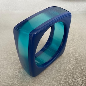 May include: A square-shaped bangle bracelet crafted from translucent resin, featuring a bold color block design with deep navy blue edges and a vibrant turquoise center section that creates a striking contrast in light and transparency.