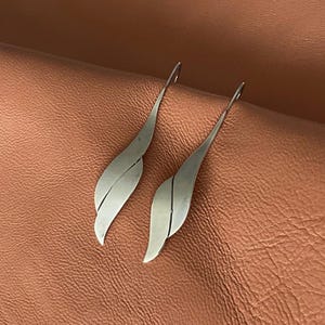 May include: A pair of silver-toned leaf-shaped earrings. Each earring has a curved, elongated design with a subtle crease down the center. The earrings are displayed on a textured, brown surface.