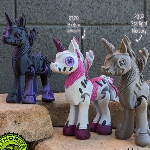 May include: Three 3D printed unicorn figurines in different colours: black with purple accents, white with pink accents, and grey. The unicorns are articulated and have a steampunk aesthetic. The text "we are paintable! kits available in the Craft Supplies section" is printed below the figurines.