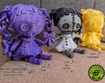 May include: Three 3D printed doll figures, one purple, one grey, and one yellow. The dolls are all in the style of rag dolls with stitched features and button eyes. The dolls are sitting on a brown surface. The text "we are paintable! kits available in the Craft Supplies section" is visible at the bottom of the image. The dolls are labeled with their color and number: 2248 Purple, 2230 Grey with paint, and 2332 Lemon Yellow.